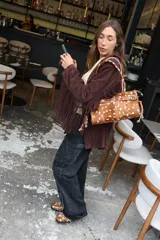 Cartera de tamaño medio confeccionada en cuero vacuno con textura de pelo y estampado animal print tipo vaca. Presenta estructura rígida, doble asa de hombro, cierre superior, detalle de tira horizontal con tachas y hebillas metálicas, y lazos decorativos en los laterales.