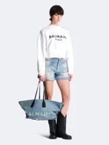 Bolso tote Balmain confeccionado en denim azul con logo estampado en blanco. Tiene doble asa corta de cuero negro y cordones laterales ajustables.