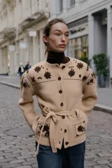 Chaqueta de punto confeccionada en mezcla de lana y alpaca, color beige. Presenta cuello redondo, manga larga, cierre frontal con botones y cinturón ajustable del mismo tejido. Incluye bolsillos delanteros de plastrón y detalles de flores bordadas en color marrón a contraste en la parte superior y en los bolsillos.