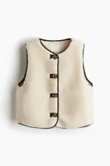 Chaleco de efecto peludito color beige, con cuello redondo, cierre frontal de botones y ribetes en contraste marrón oscuro. Incluye bolsillos laterales discretos y forro interior.