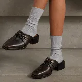 Zueco de cuero vacuno color marrón oscuro con detalle de nudo en el empeine, punta cuadrada y taco bajo de madera.