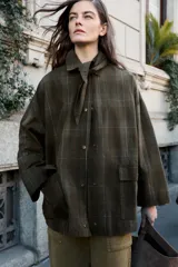 Chaqueta de algodón encerado con diseño de cuadros en tonos verde oliva, beige y líneas rojas. Presenta cuello solapa, cierre frontal con botones a presión metálicos, bolsillos de plastrón con solapa y un detalle de pañuelo desmontable sobre los hombros.