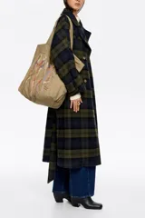 Bolso tipo shopper de color beige, con diseño amplio y doble asa de hombro. Presenta un detalle frontal de bordados coloridos con hilos colgantes que forman un diseño artístico.