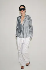 Camisa de cuello mao y manga larga con puños anchos, de color gris satinado. Cuenta con un bolsillo de plastrón en el pecho con detalles bordados a tono, al igual que los puños, y cierre frontal con botones ocultos bajo solapa.