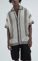 Camisa de manga corta con tejido de punto abierto tipo crochet, diseño oversize con cuello solapa y cierre frontal de botones. Presenta un patrón geométrico en color beige con detalles de bordes en negro.