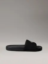 Ojotas tipo slide Calvin Klein Jeans, color negro, con banda superior de lona que presenta el logo de la marca bordado en blanco. Posee suela de EVA con plantilla ergonómica moldeada y el nombre de la marca grabado en el lateral.