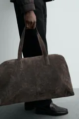 Bolso de viaje de gran tamaño confeccionado en piel con acabado de serraje. Cuenta con un compartimento principal con cierre de cremallera e imanes, interior con bolsillo con cierre y bolsillo plano, y dos asas de mano de piel.