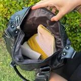 Bolso negro de cuero sintético con doble asa de hombro y correas decorativas frontales con hebillas metálicas. Incluye cierre superior y bolsillo interior.