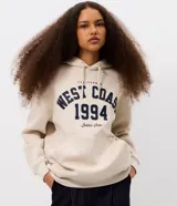 Buzo canguro de corte oversized color beige, con capucha ajustable mediante cordones y bolsillo frontal. Presenta una estampa estilo college en el pecho con la inscripción 'West Coast 1994' en color azul marino.