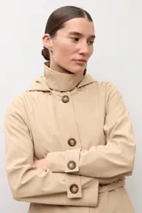 Trench clásico color beige, con cuello alto, capucha desmontable, cierre frontal de botones marrones, bolsillos ribeteados y cinturón ajustable con hebilla.