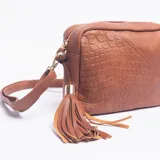 Bandolera de tamaño mediano color marrón, con diseño combinado de textura lisa y efecto croco en uno de sus laterales. Cuenta con correa larga ajustable, cierre superior y detalle de borla decorativa en uno de los costados.