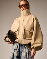 Chaqueta color beige de estilo utilitario con cuello alto y cierre frontal parcial. Presenta mangas amplias con puños elásticos y dos bolsillos frontales tipo cargo con solapa.