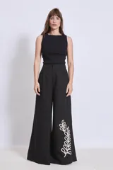 Pantalón negro de corte palazzo con bolsillos laterales y apliques bordados de hojas en color crudo en una pierna.