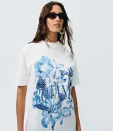 Remera de algodón blanca de corte largo, manga corta y cuello redondo. Presenta un gran estampado floral en tonos azules con la inscripción "Botanist" en el frente.