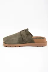 Zueco de gamuza color verde oliva, con diseño cerrado en la parte frontal y hebilla metálica dorada ajustable en el empeine. Presenta una suela de goma dentada en color marrón.