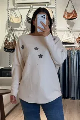 Buzo tejido de punto en color beige, con diseño de flores aplicadas en tono gris oscuro y detalles de brillos plateados que irradian desde las flores. Presenta cuello redondo amplio, mangas largas con caída holgada y corte recto.