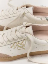 Championes de cuero color blanco con puntera y suela de goma marrón claro. Presentan detalles bordados dorados en los laterales y cordones anchos en color crudo.