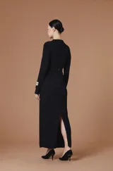 Vestido largo entallado de color negro, con cuello alto y mangas largas. Presenta una textura plisada con ondas verticales y un cinturón integrado con hebilla metálica dorada en la cintura.