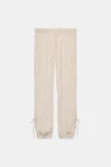 Pantalón de mujer color beige claro, de tiro medio y calce holgado, confeccionado en tejido ligero y fluido. Presenta cintura elástica ajustable con lazada, bolsillos laterales y aberturas ajustables con lazadas en las piernas que terminan en bajo abullonado con elástico.