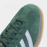 Championes Adidas Gazelle Indoor de gamuza verde oscuro, con las tres tiras características en color blanco y el nombre del modelo en letras doradas en el lateral. Poseen suela de goma translúcida y detalles en color celeste claro en el talón.