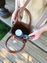 Bolso matera de cuero color marrón con doble asa de mano y correa larga desmontable. Incluye accesorio decorativo de borlas rosas y blancas.