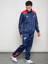 Conjunto deportivo Umbro del Club Nacional de Football, compuesto por una campera con cierre frontal y pantalón a juego. La campera es de color azul con detalles en rojo en los hombros y líneas blancas, presentando el logo de la marca y el escudo del club en el pecho. El conjunto está confeccionado en tejido sintético con acabado satinado.