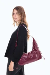 Cartera tipo hobo de material sintético con acabado brillante, color bordó. Cuenta con cierre superior de cremallera, correa ajustable y detalles de cordones decorativos en los laterales.