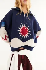 Sweater de punto de algodón con cuello polo y cierre frontal. Presenta un diseño con intarsia de sol y montañas en tonos azul, blanco y rojo, con detalle de parche en la manga.