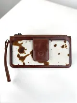 Billetera tipo porta celular de cuero, con diseño combinado en cuero liso marrón y cuero con pelo vacuno estampado. Cuenta con cierre superior, correa de muñeca y solapa frontal con cierre magnético y logo de búho grabado.