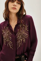 Camisa de viscosa color violeta con cuello clásico, manga larga y cierre frontal con botones. Presenta un diseño bordado con lentejuelas doradas en ambos lados del pecho.
