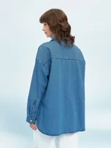 Camisa de denim azul de corte oversize, con mangas largas, cuello clásico, un bolsillo aplicado en el pecho y cierre frontal con botones metálicos.