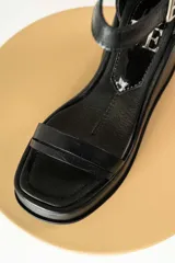 Sandalias negras de cuero sintético con plataforma y taco de cuña. Tienen dos tiras en el empeine y una tira con hebilla alrededor del tobillo.