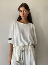 Remera básica de corte oversize y diseño cropped, color crudo, con mangas cortas y etiqueta decorativa en la manga derecha.