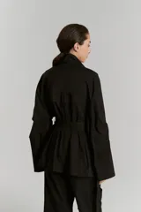 Parka negra de gabardina con corte amplio, cuello camisero, manga amplia con recortes y presillas en puños, cartera de botones oculta, bolsillos plaque con tapa, canesú con pestaña en delantero y cinturón con medialuna metálica.