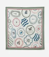 Pañuelo de satén con estampado tropical que incluye palmeras, casas, olas y otros íconos que recuerdan a las brasilidades. Presenta un borde verde y detalles en rojo, azul y rosa.