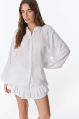 Camisa blanca de lino con textura arrugada, cuello mao, botones frontales y mangas largas abullonadas con puños elásticos. Presenta detalles de encaje calado en los hombros.