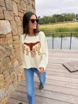 Sweater de punto fino color crema, con cuello redondo en tono contraste. Presenta un diseño frontal artesanal con figura de búfalo en cuero y detalles bordados en relieve.