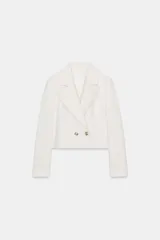 Blazer corto blanco de cuello solapa y manga larga con hombreras. Presenta un cierre frontal cruzado con dos botones dorados.