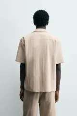 Camisa de manga corta y corte relajado, confeccionada en tejido de algodón con estructura de rayas verticales. Presenta cuello bowling, cierre frontal con botones y aberturas laterales en el bajo.
