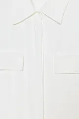 Camisa de cuello solapa y manga corta, confeccionada en tejido satinado. Presenta falsos bolsillos con solapa en el delantero, bajo asimétrico y cierre frontal con botones.