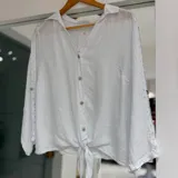 Camisa blanca de lino con mangas largas abullonadas, cuello camisero, cierre frontal con botones y detalle de nudo en el dobladillo delantero.