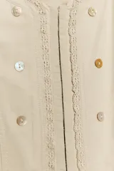 Chaqueta de cuello redondo y manga larga en color beige. Presenta un diseño con doble hilera de botones decorativos en el frente, detalles de encaje a tono y cierre frontal mediante ganchos metálicos ocultos.