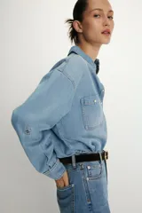 Camisa de denim de corte oversize con cuello solapa, manga larga con puños, cierre frontal con botones a presión y bolsillos de plastrón en el delantero.