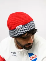 Gorro tipo beanie de punto acanalado en color azul marino, con vuelta en color rojo y blanco a rayas. Incluye etiqueta lateral con el logo de Umbro y el escudo del Club Nacional de Football.