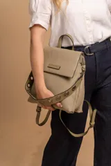 Mochila tipo cartera con tapa, confeccionada en símil cuero color suela. Cuenta con doble compartimento, división interna, cierre de seguridad y bolsillo trasero con cierre. Incluye una correa frontal decorativa con tachas metálicas.