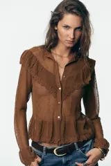 Chaqueta de punto pointelle color tostado, con cuello subido y manga larga. Presenta detalles de volantes en el pecho y puños, y cierre frontal con botones.
