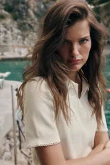 Top de punto color crudo, con cuello polo y escote en V. Cuenta con mangas cortas y detalle de puntilla con forma de onda en el cuello y los puños.