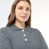 Sweater de punto gris con cuello polo, manga larga y puños acanalados. Presenta una abertura frontal con tres botones decorativos tipo joya con brillos.