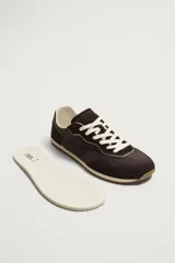 Zapatilla deportiva estilo barefoot, color marrón, con diseño de paneles en combinación de materiales y detalles en serraje. Cuenta con puntera redondeada y ancha, cordones en color crema y suela fina de goma flexible en contraste.