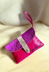 Sobre de mano Carolina de Cunto modelo Saria de la línea Ottenta, confeccionado en cuero fucsia con solapa de cuero metalizado texturizado y detalle de tira dorada. Cuenta con correa de mano y cierre con broche imán.